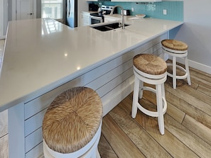 A modern kitchen with a large island, two stools with woven seats, and a blue tiled backsplash. - Copano Breeze by Miss Kitty’s (Rockport)