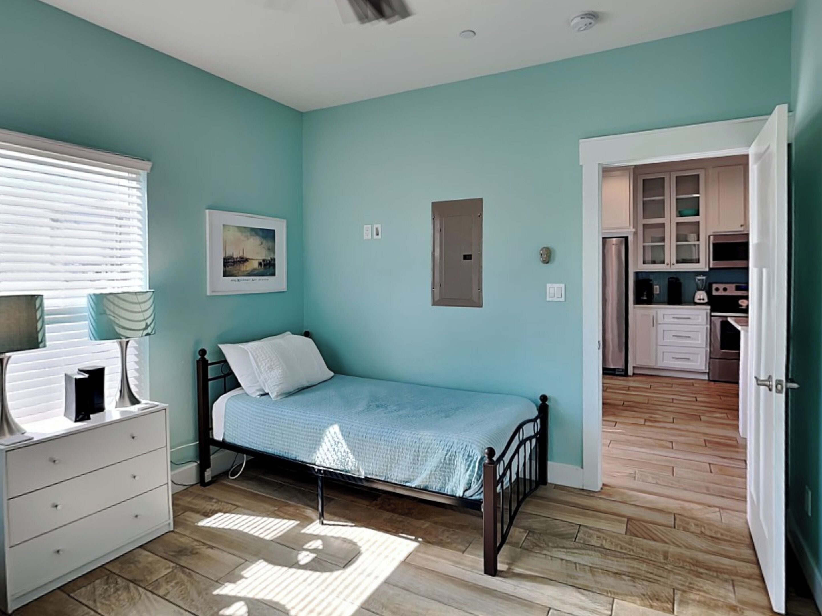 Un dormitorio con cama, cómoda, ventana con persianas, un cuadro en la pared y una puerta que da a una cocina.