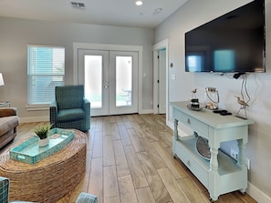 A living room with a green armchair, a wicker ottoman, a light blue console table with decorative items, and a flat-screen TV mounted on the wall. - Copano Breeze by Miss Kitty’s (Rockport)