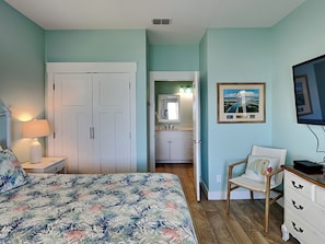 A bedroom with a bed, a chair, a dresser, a TV, and a bathroom visible through an open door.