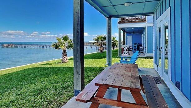 A covered outdoor area with a wooden picnic table, blue chairs, and a view of the ocean and pier. - Copano Breeze by Miss Kitty’s (Rockport)