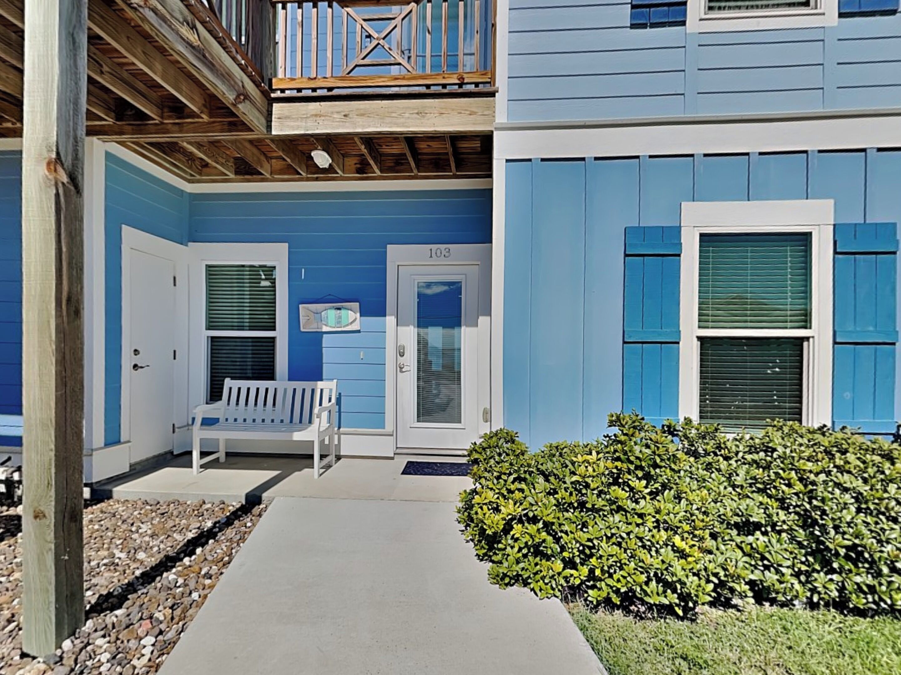 A coastal building with a blue exterior, a wooden balcony, a bench, and a hedge.
