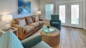 A living room with a brown sofa, a blue patterned sofa, a wicker coffee table, and a teal armchair. There is a painting of a sea turtle on the wall and a window with blinds. - Copano Breeze by Miss Kitty’s (Rockport)