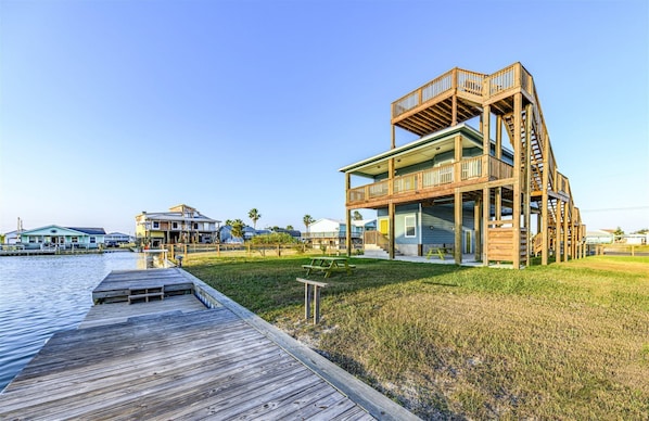 A waterfront property with a wooden deck, a multi-level house with a balcony, and a grassy area.