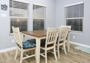 Uma sala de jantar com uma mesa de madeira e quatro cadeiras, três janelas com persianas e um tapete estampado no chão.