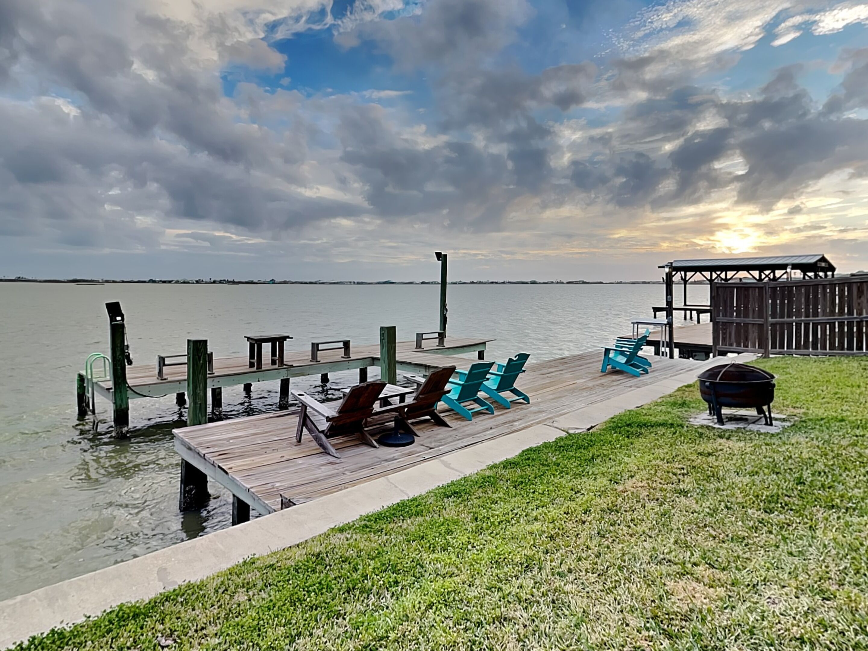 A wooden dock extending into a body of water with chairs and a grill, and a covered structure in the background.