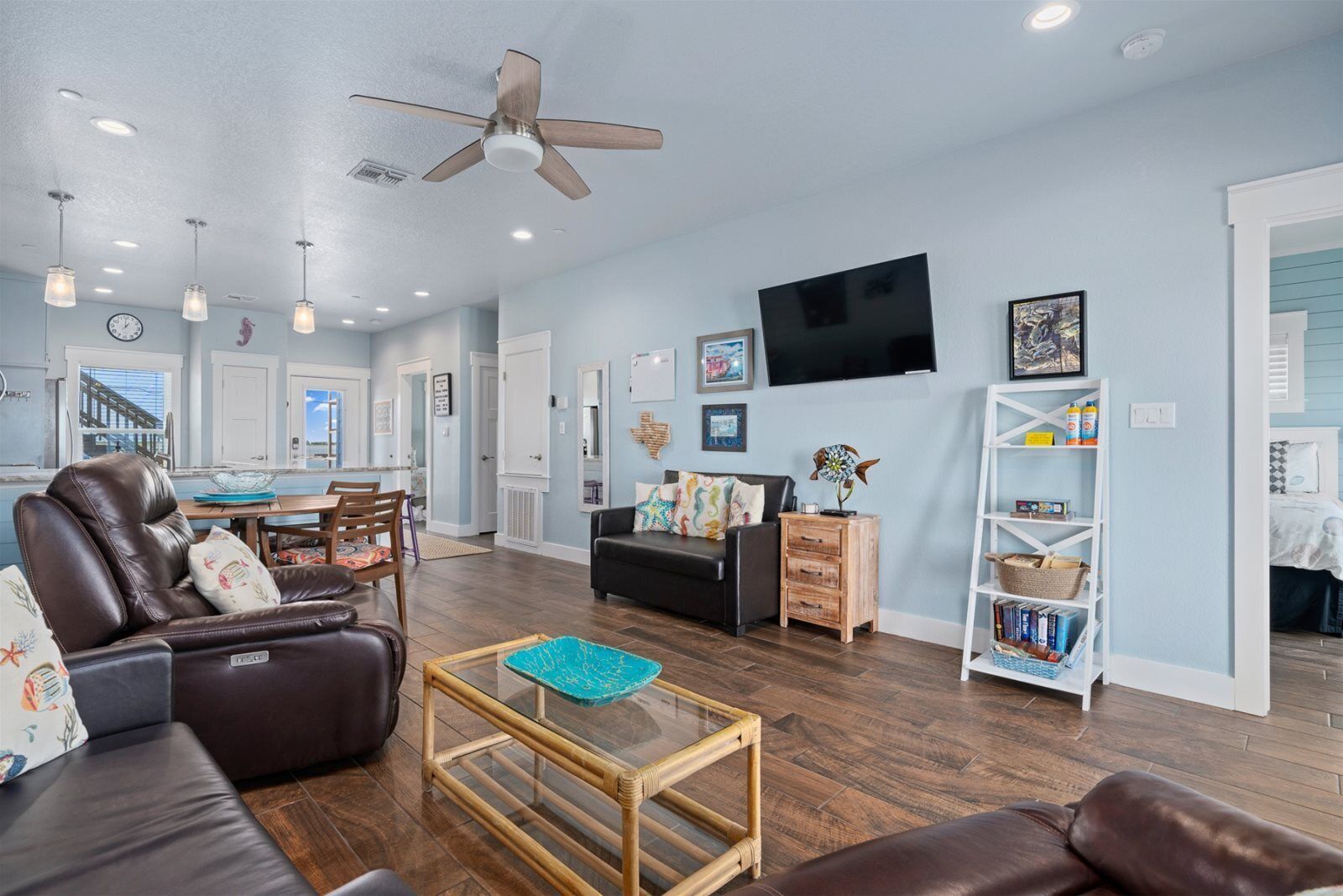 A spacious living room with a dark brown leather sofa, a glass coffee table, and a wooden side table. A ceiling fan and recessed lighting are visible.