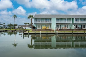 A two-story building with a balcony, a dock, and a calm water body in front. - Coastal Bliss by Miss Kitty’s (Rockport)