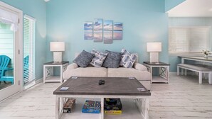 Ein Wohnzimmer mit einer türkisfarbenen Wand, einem Sofa mit gemusterten Kissen, einem Couchtisch mit Fernbedienung und einem Bücherregal mit Gesellschaftsspielen.