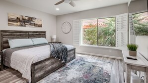 A bedroom with a wooden bed, a ceiling fan, a large window with shutters, and a view of greenery outside. - The Perfect Tucson Townhome in Catalina Foothills (Tucson)