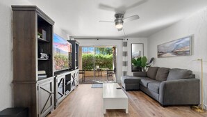 A modern living room with a grey sectional sofa, a white coffee table, and a TV on a wooden cabinet. There is a ceiling fan, a floor lamp, and a large window with a view of greenery. - Lovely 2-Bedroom Apartment in Scottsdale! (Scottsdale)