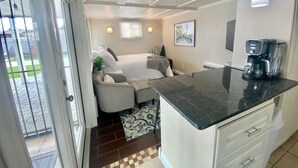 Habitació compacta amb llit, zona de seients petita, cuina americana amb cafetera i vista per una porta de vidre.