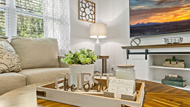 Una sala de estar con un sofá, una mesa de centro, una bandeja decorativa con las palabras "be" y "our", una planta en maceta, una lámpara y un cuadro de un atardecer.