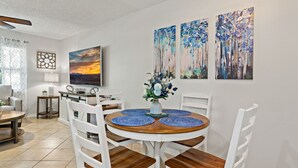 Un comedor con una mesa redonda de madera, sillas blancas y manteles individuales azules. Hay tres cuadros en la pared, una lámpara y una ventana con cortina.
