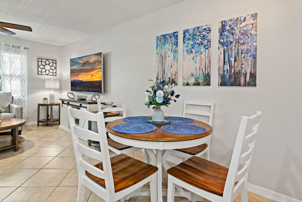 A dining area with a round wooden table, white chairs, and blue placemats. There are three paintings on the wall, a lamp, and a window with a curtain.