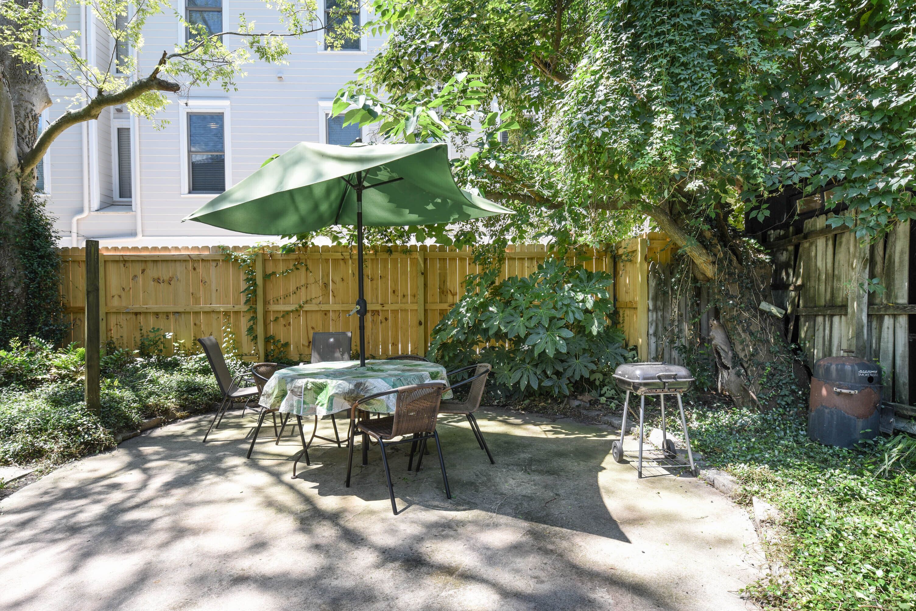 Une terrasse avec une table et des chaises sous un parasol vert, entourée d’une clôture en bois et de verdure.