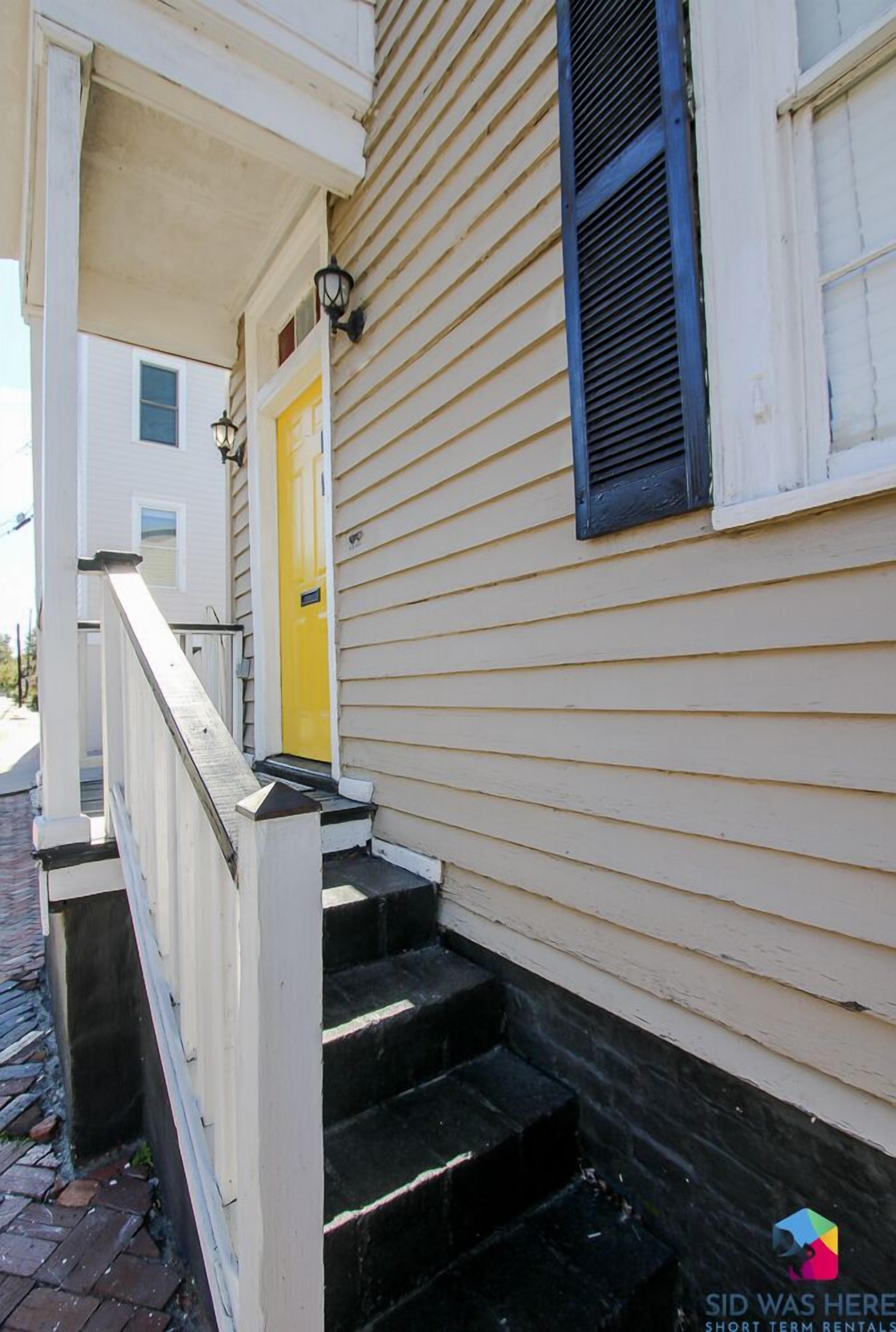 A two-story house with a yellow front door, a black window shutter, and a white railing on the front steps.