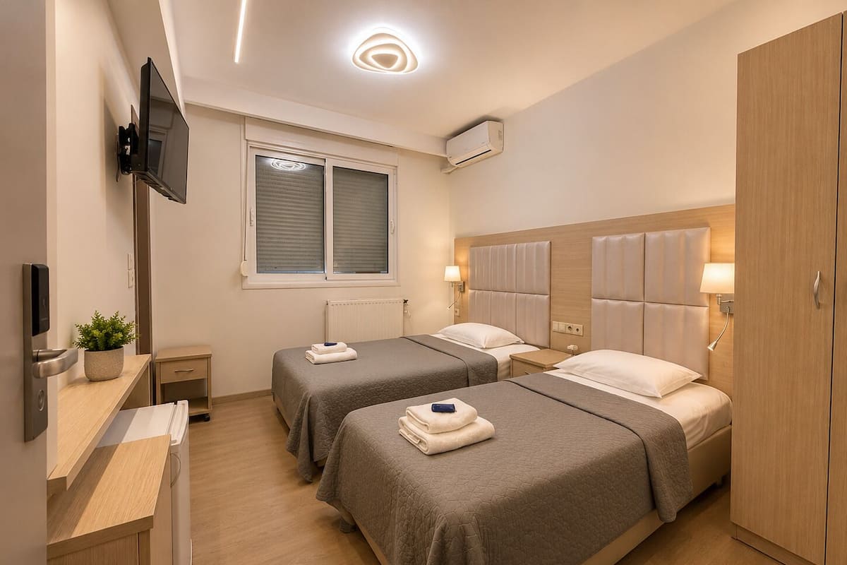 A hotel room with two beds, a TV, a desk, a plant, and a window with blinds.