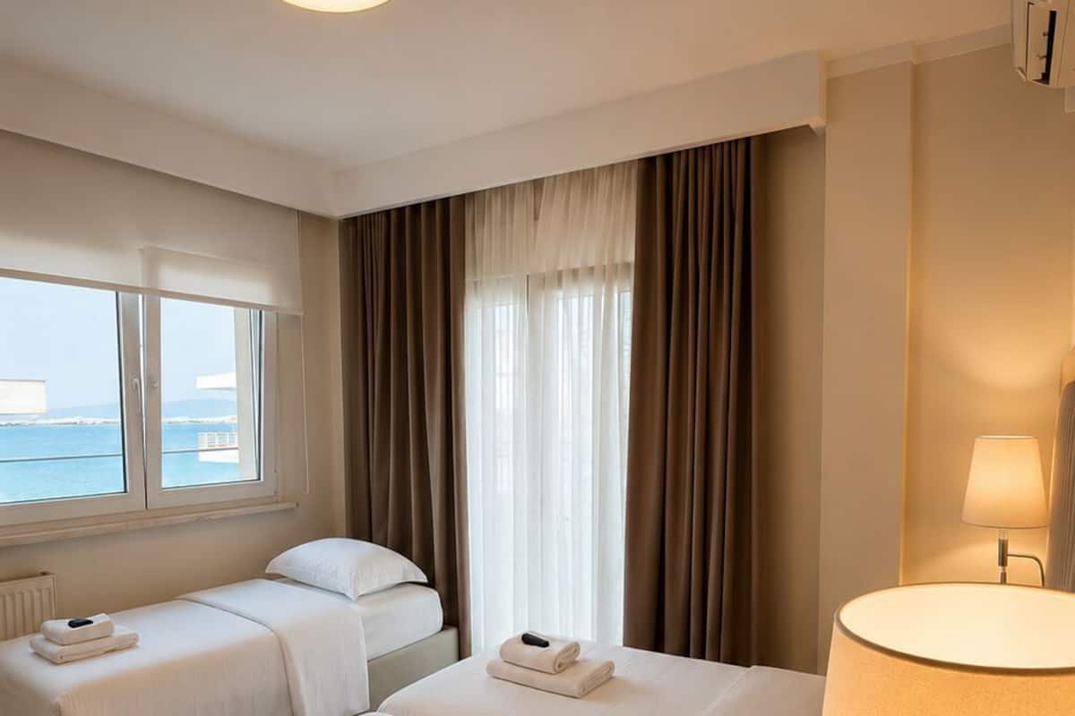 A hotel room with two beds, a window with a view, a lamp, and a radiator.