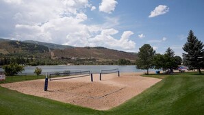 A beach volleyball court with a net, surrounded by a grassy area and trees, overlooking a lake with hills in the background.