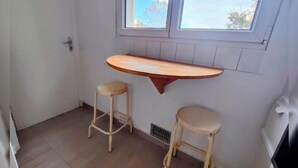 Ein kleiner Raum mit zwei hölzernen Hocker, einer Holzablage und einem Fenster mit Blick auf Bäume.