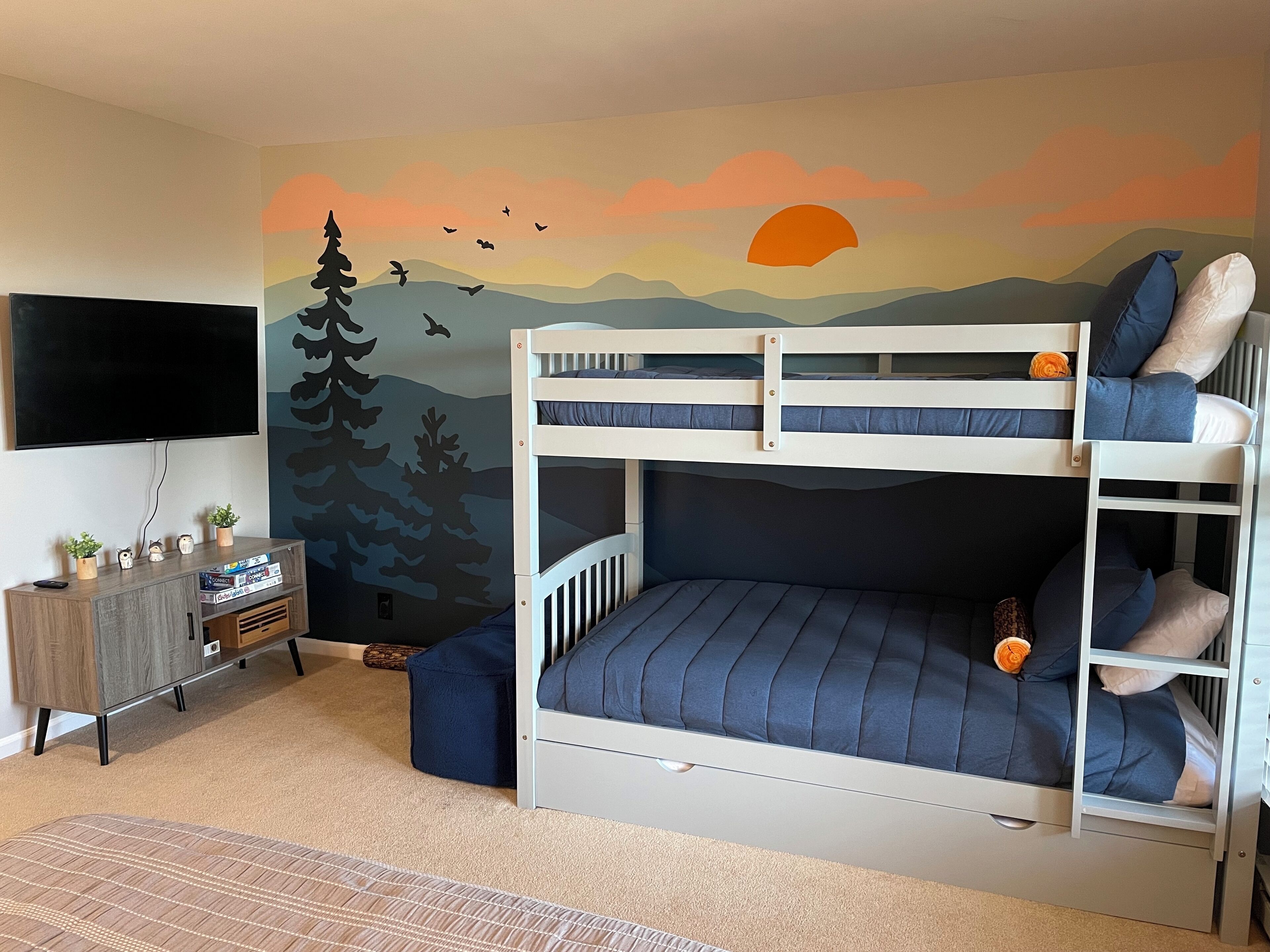 Une chambre avec un lit superposé, ornée d’une fresque représentant un coucher de soleil et des montagnes, ainsi qu’une télévision et un petit meuble agrémenté de plantes.
