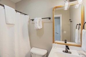 A bathroom with a white toilet, a towel rack with a towel, a shower curtain, and a large mirror with lights.