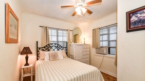 A bedroom with a bed, a nightstand with a lamp, a dresser, a ceiling fan, and a window with blinds.