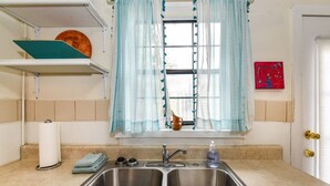 A kitchen with a double sink, a window with blue curtains, a shelf with a basket, and a red artwork on the wall.