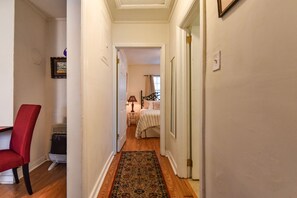 A narrow hallway with a wooden floor, a red chair, and a bed in the background.