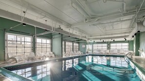 Una amplia piscina interior con paredes de vidrio transparente, múltiples ventanales y sillones blancos alrededor del borde.