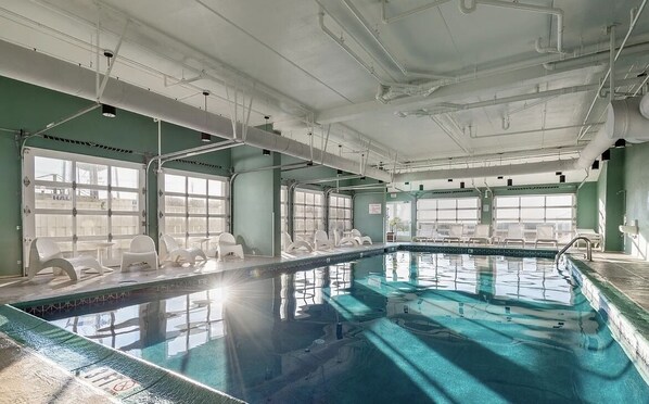 Une grande piscine intérieure aux parois en verre transparent, dotée de nombreuses fenêtres et entourée de chaises longues blanches.