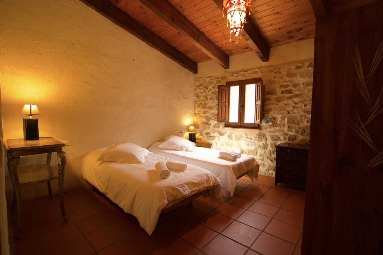 Une chambre avec deux lits, un mur en pierre, des poutres en bois et une fenêtre avec des volets.