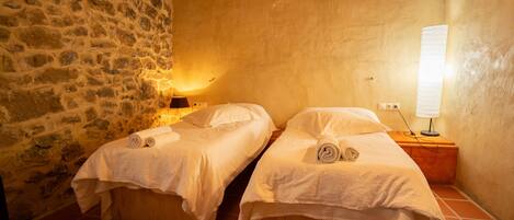 A room with two wooden beds, stone walls, and a nightstand with a lamp.