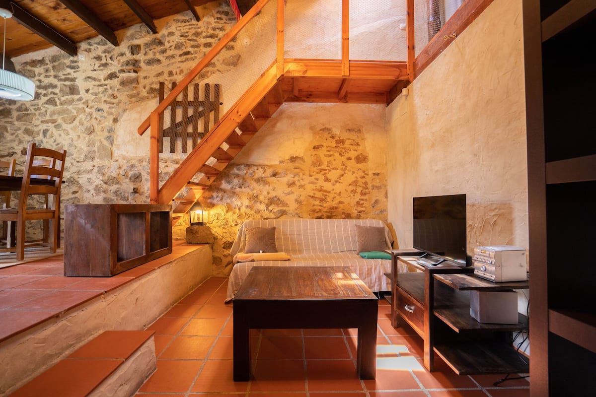 Ein gemütliches Zimmer mit Steinwand, einer Holztreppe, einer Couch, einem Holztisch und einem Fernseher.