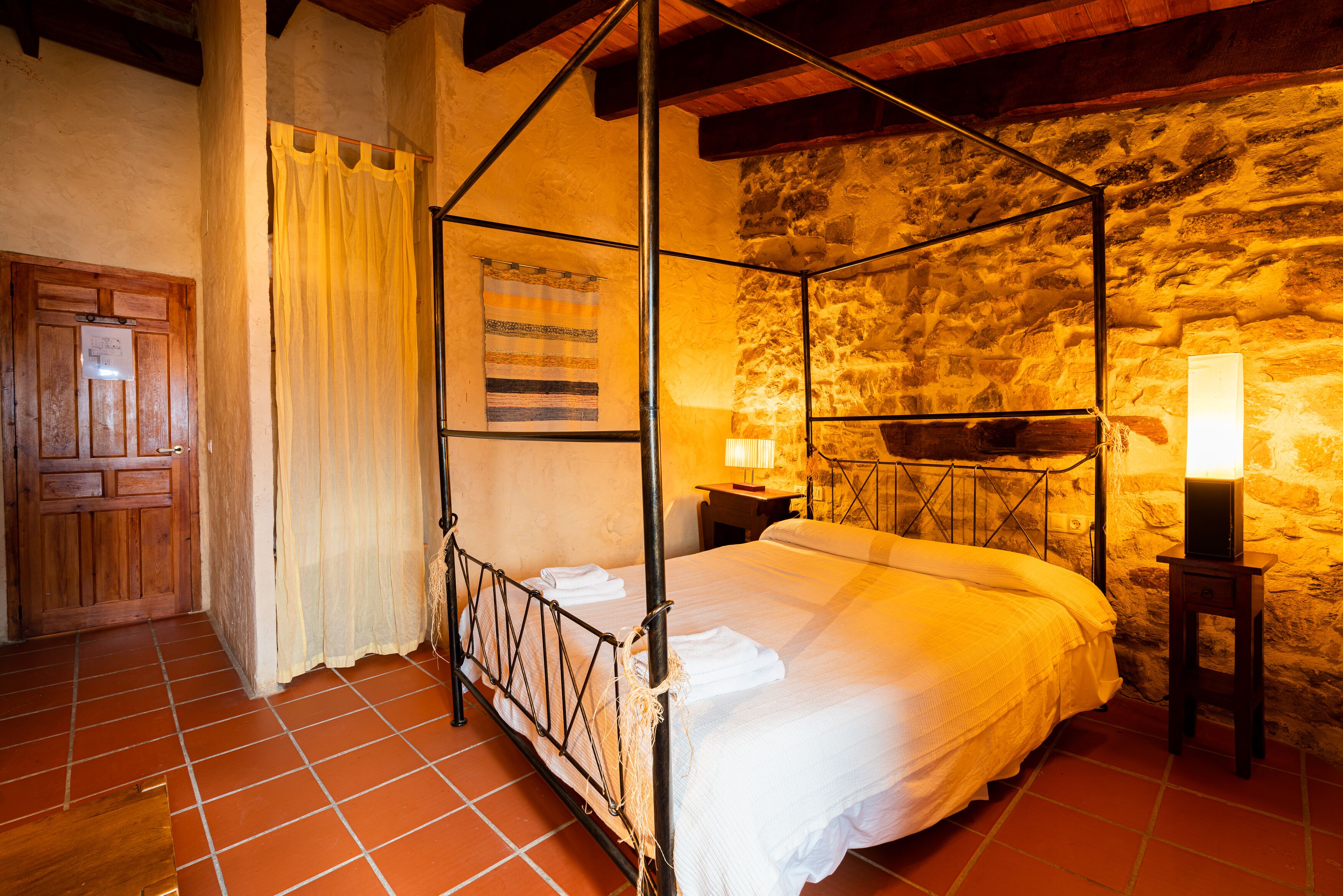 A four-poster bed with white linens, a stone wall, and a wooden door.