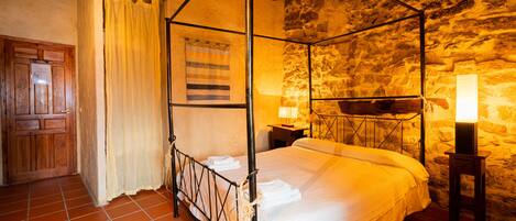A four-poster bed with white linens, a stone wall, and a wooden door.