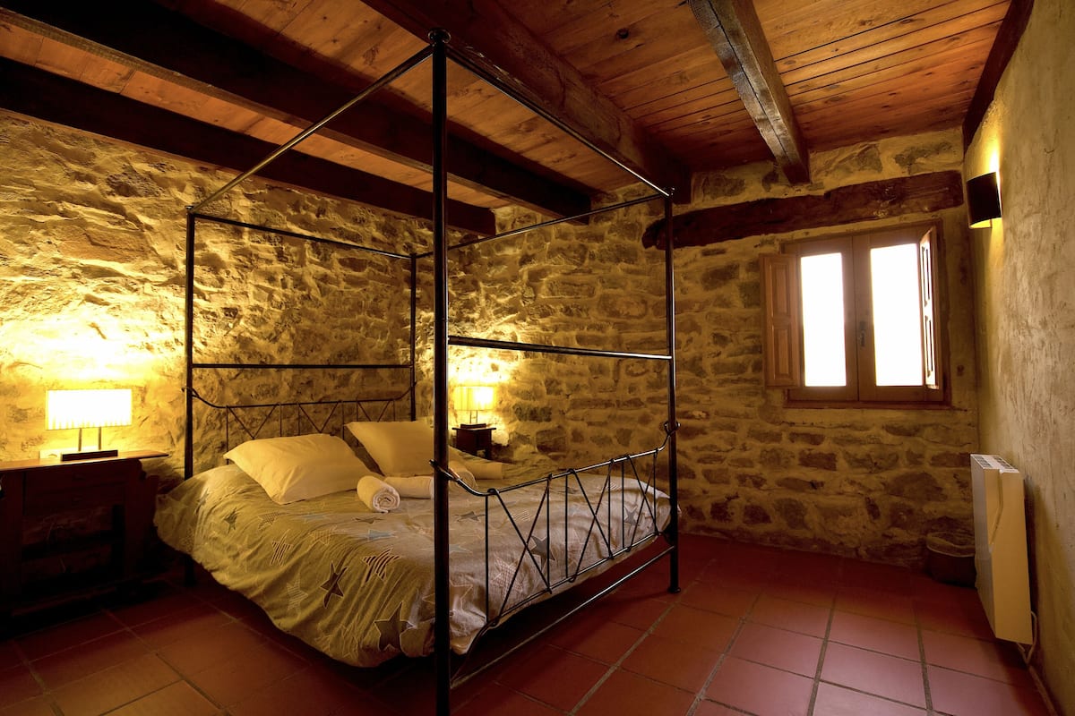Ein Bett mit Baldachin in einem rustikalen Zimmer mit Steinwänden, Holzbalkendecke und einem Fenster, das für natürliches Licht sorgt.