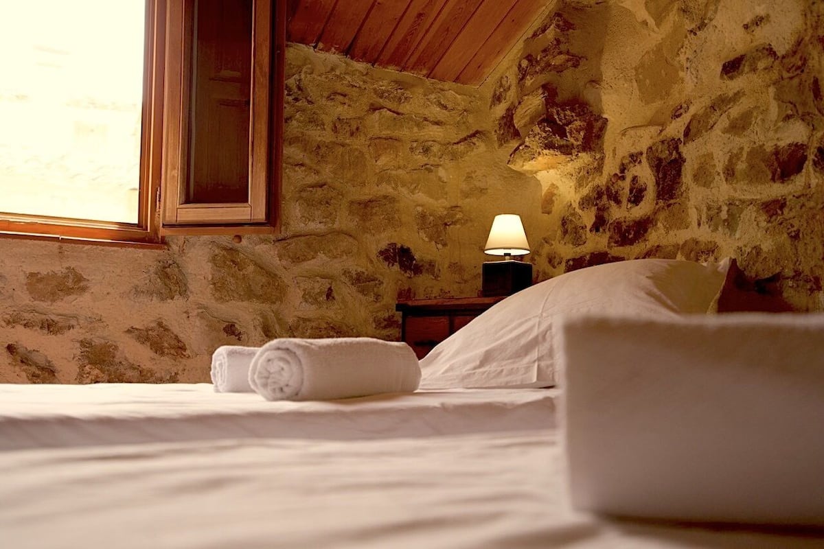 Ein Schlafzimmer mit Steinwänden, Holzbalkendecke, einem Bett mit weißen Leinentüchern, einem zusammengerollten Handtuch, einem Nachttisch mit Lampe und einem Fenster mit Tageslicht.