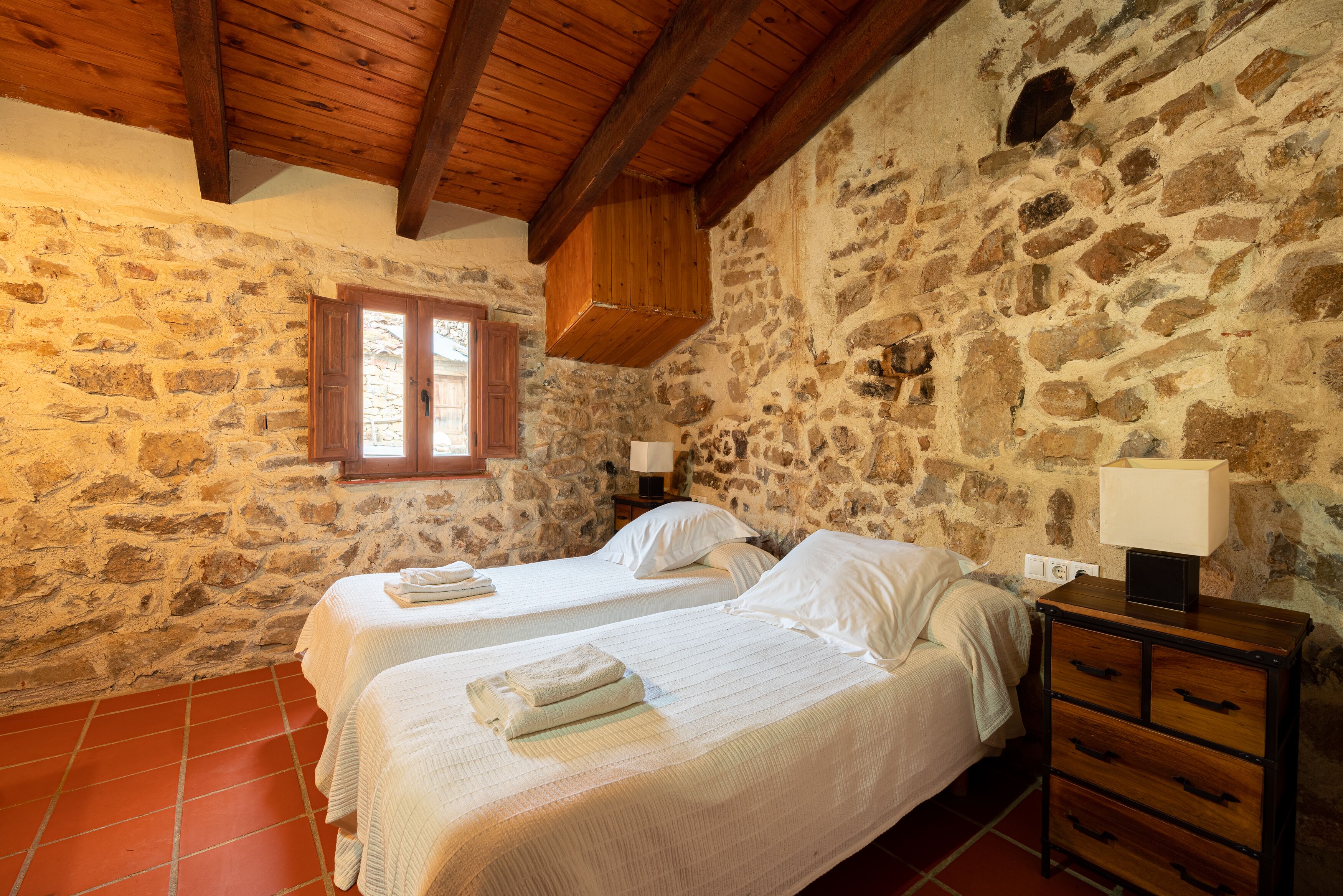 A room with two beds, wooden beams, stone walls, a window, and a nightstand with a lamp.