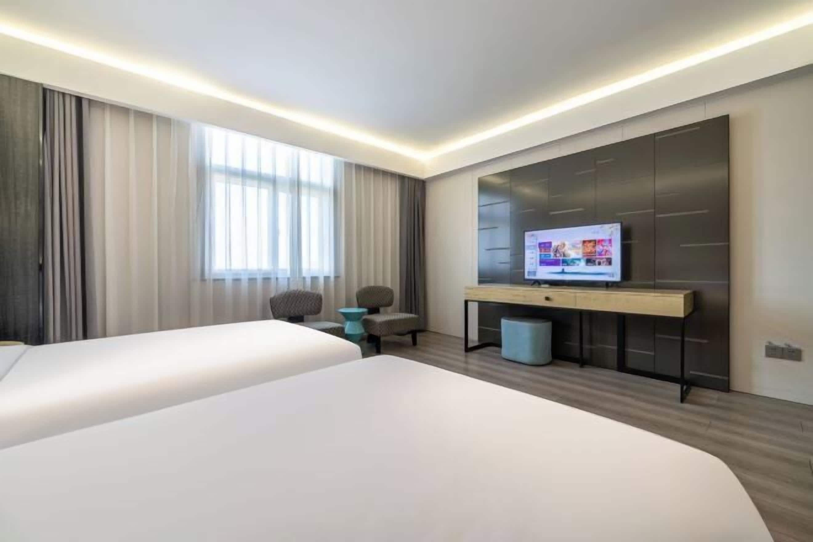 A hotel room with two beds, a TV, a desk, and a large window with curtains.