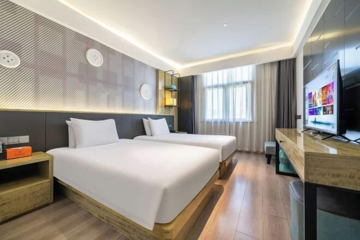 A hotel room with two beds, a desk with a TV, a clock on the wall, and a window with curtains.