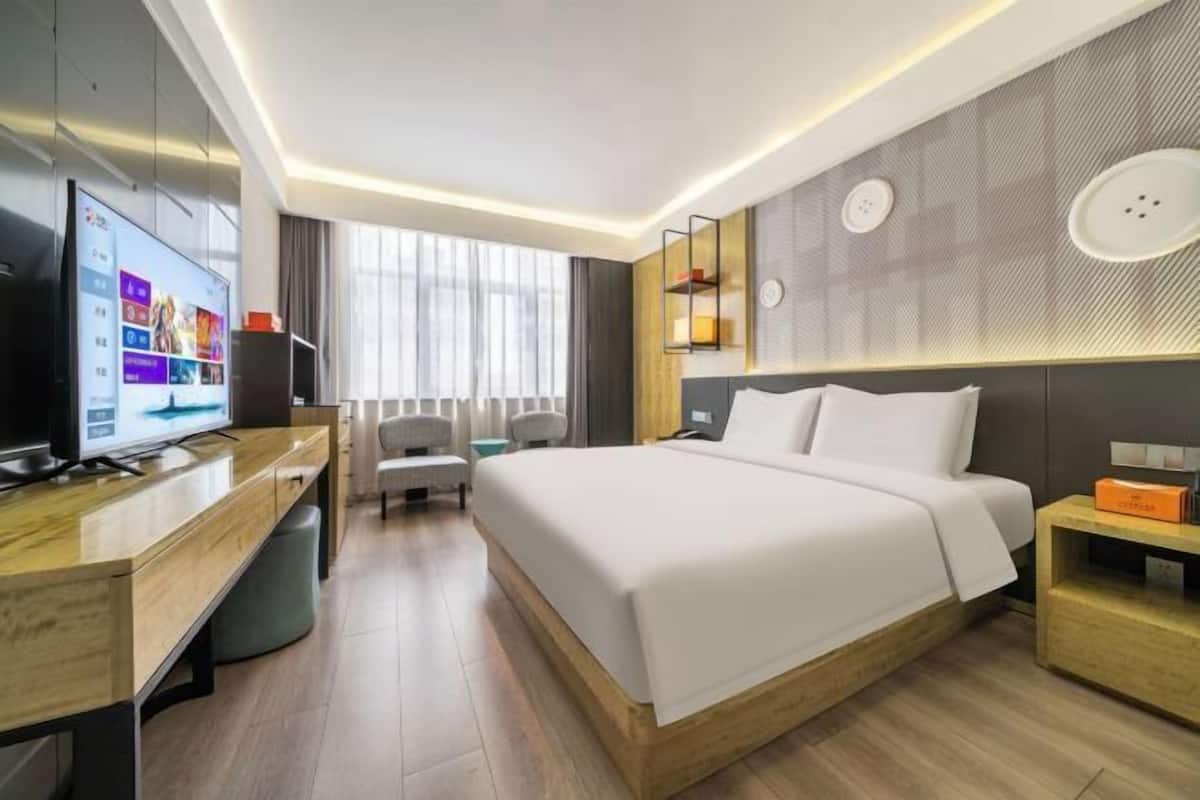 A modern hotel room with a large bed, a desk with a flat-screen TV, a seating area, and a large window.