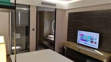 A modern hotel room with a TV on the wall, a bedside table, and a glass partition separating the bathroom.