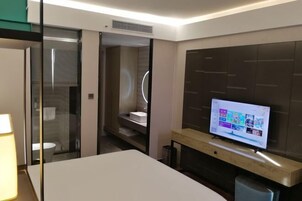 Una habitación de hotel moderna con un televisor en la pared, una mesita de noche y un separador de vidrio que divide el baño.