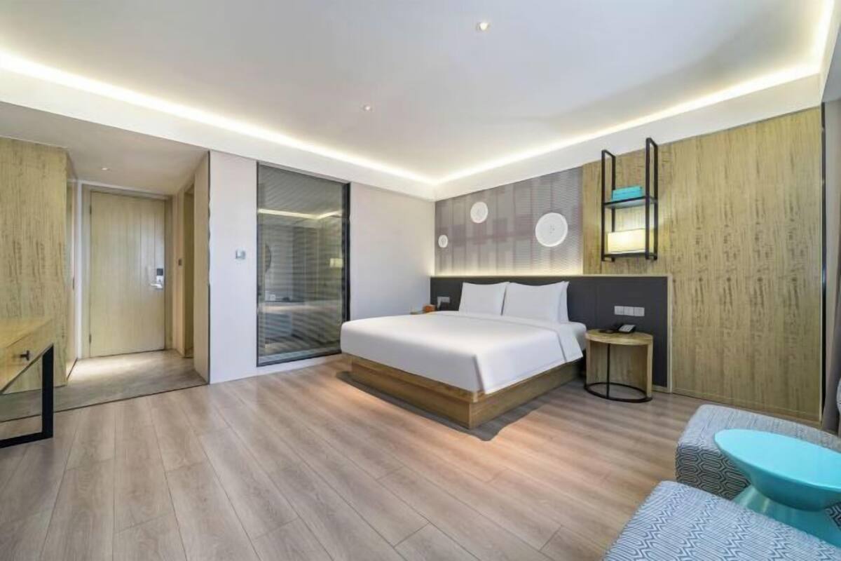 A modern hotel room with a large bed, a small bedside table, a built-in wardrobe, and a seating area with a sofa and armchair.