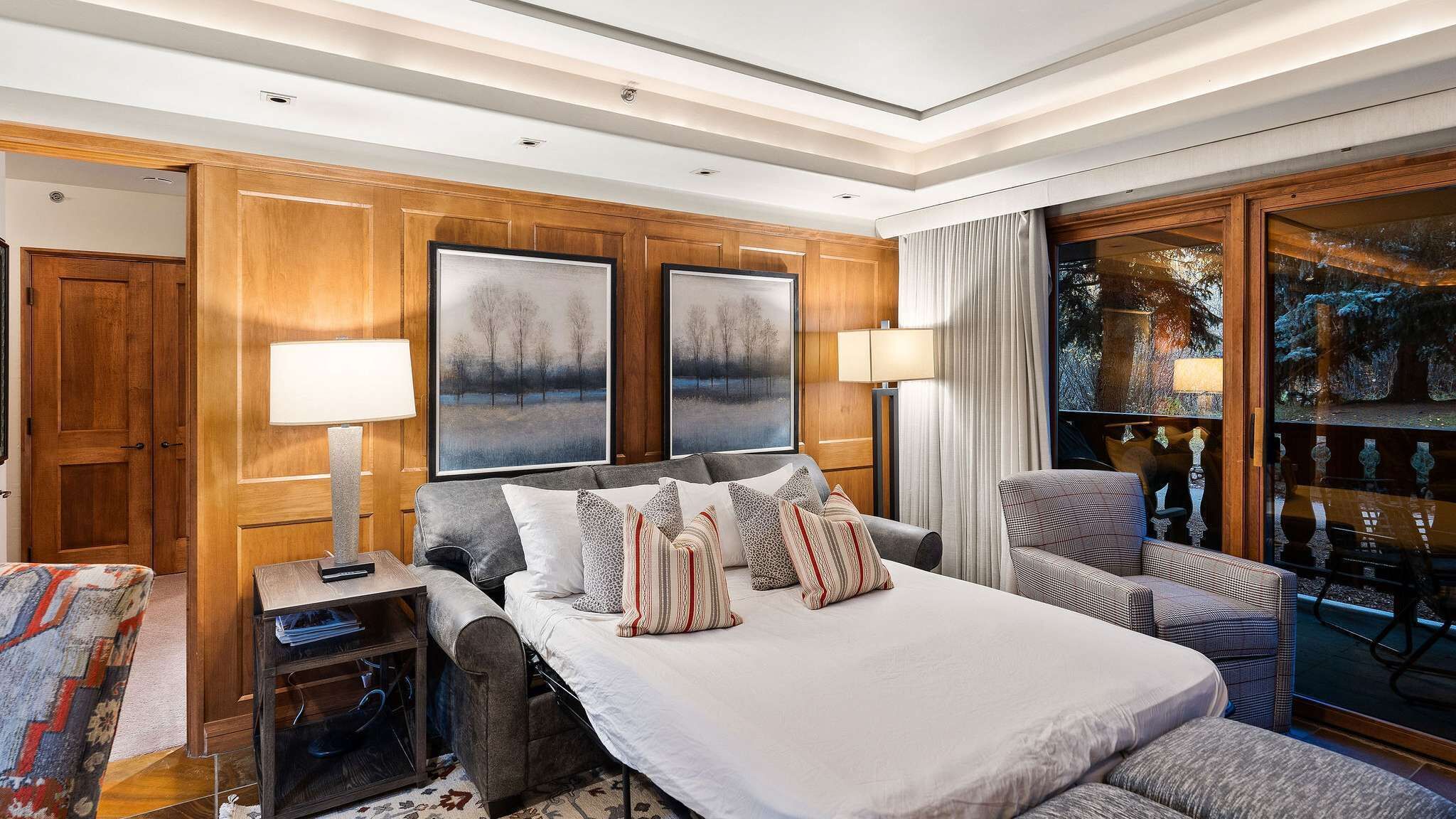 A hotel room with a large bed, two side tables, a chair, a window with a view, and wooden paneling on the walls.