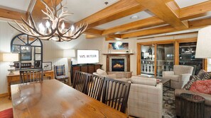 A spacious living room with a wooden dining table, comfortable seating, a fireplace, and a large TV screen displaying 'VALL BUTLER'. - VB | 6BR Slopeside Lodge - Ski In/Ski Out (Vail)