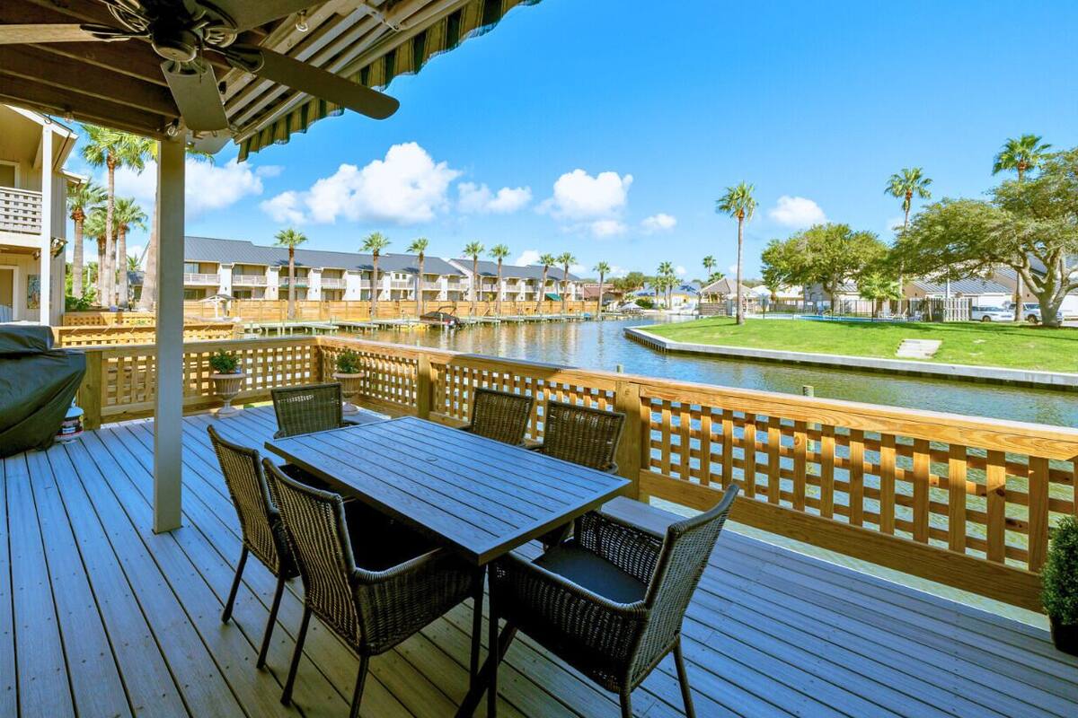 A wooden deck with a table and chairs, overlooking a pool and a lawn with palm trees.