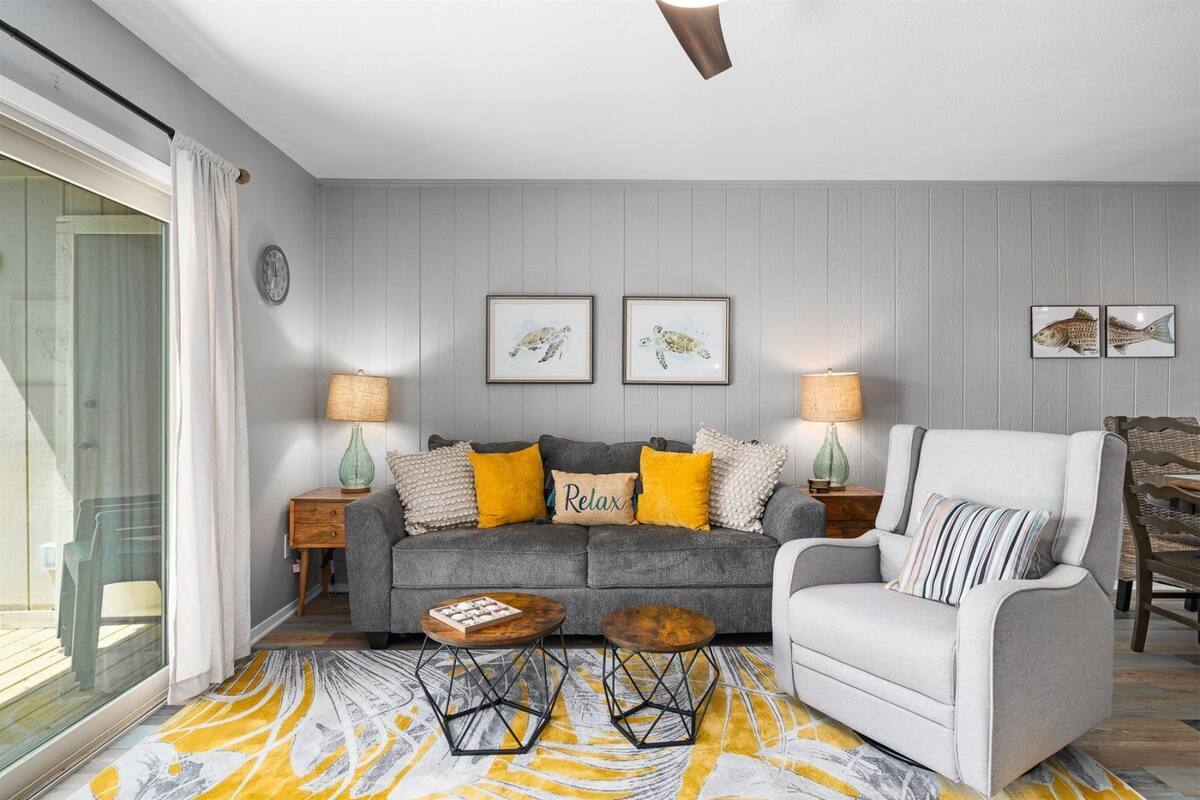 A living room with a grey sofa, a white armchair, and a coffee table. There are two lamps on side tables, a rug, and framed pictures on the wall.